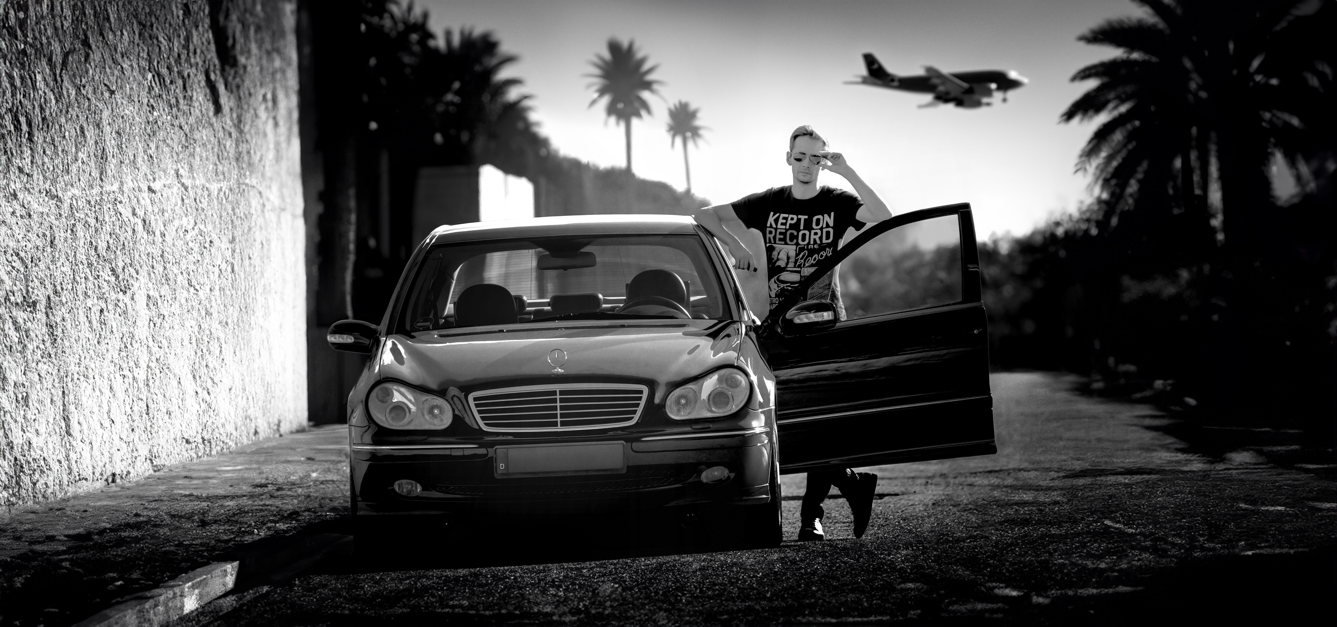 Norman Claim stands behind the open driver’s door of a Mercedes-Benz, wearing aviator sunglasses, while palm trees sway gently in the wind and an airplane passes overhead in a tropical black-and-white scene.