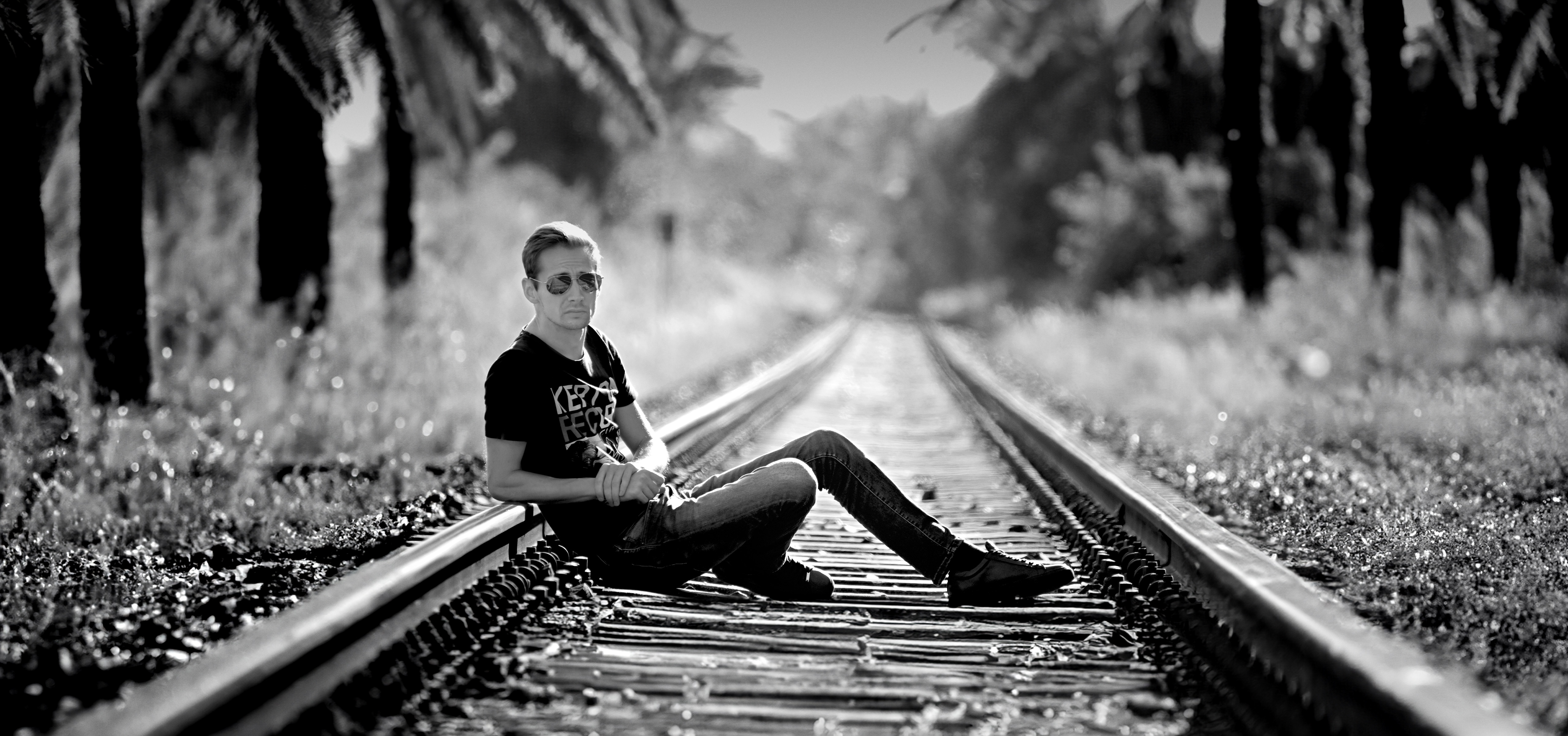 Norman Claim sits on the railroad tracks, looking directly at the camera, while a tropical palm landscape stretches along the tracks.