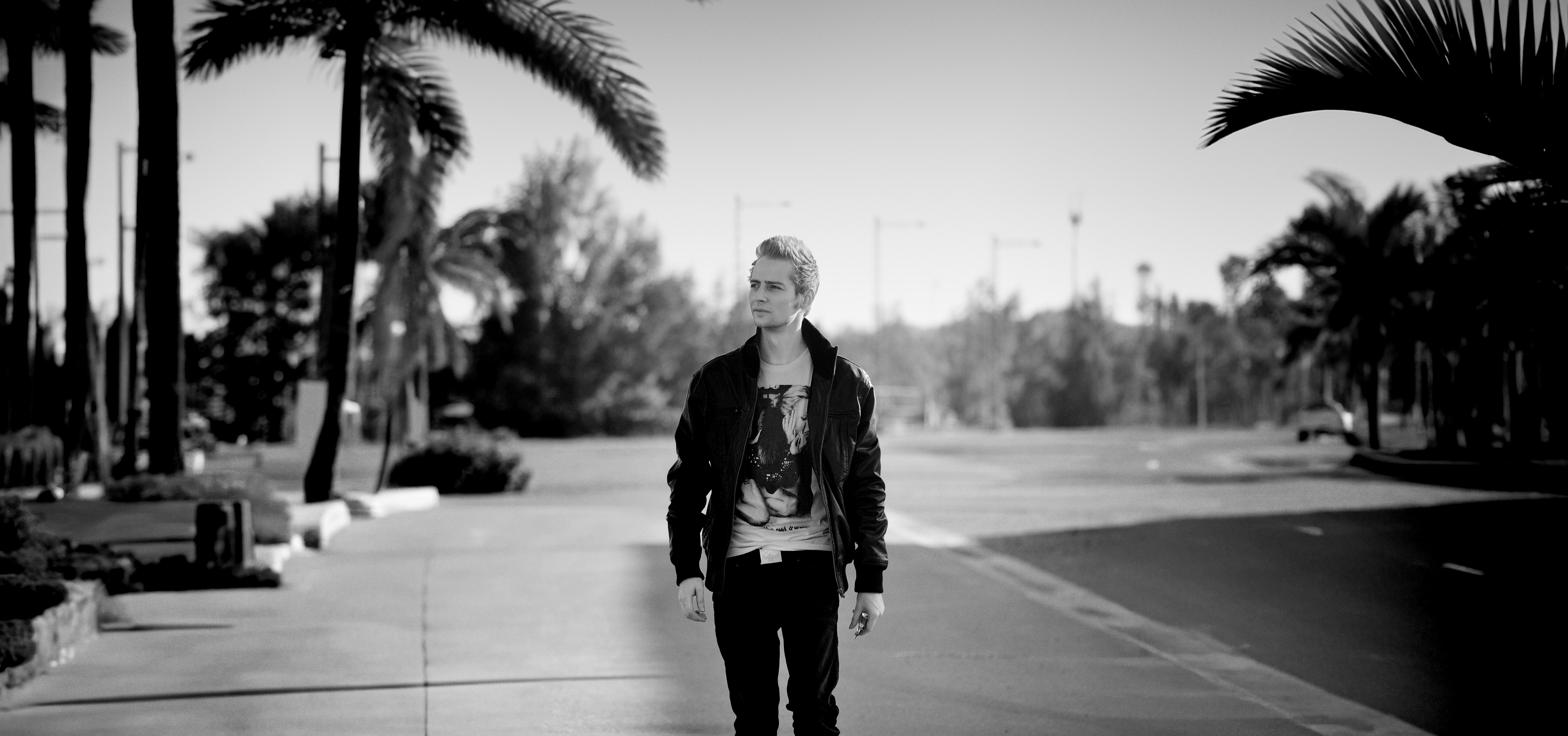 Norman Claim walking confidently along a palm-lined street, tropical trees moving in a gentle breeze, sunlight filtering through shifting shadows, black-and-white cinematic photography.